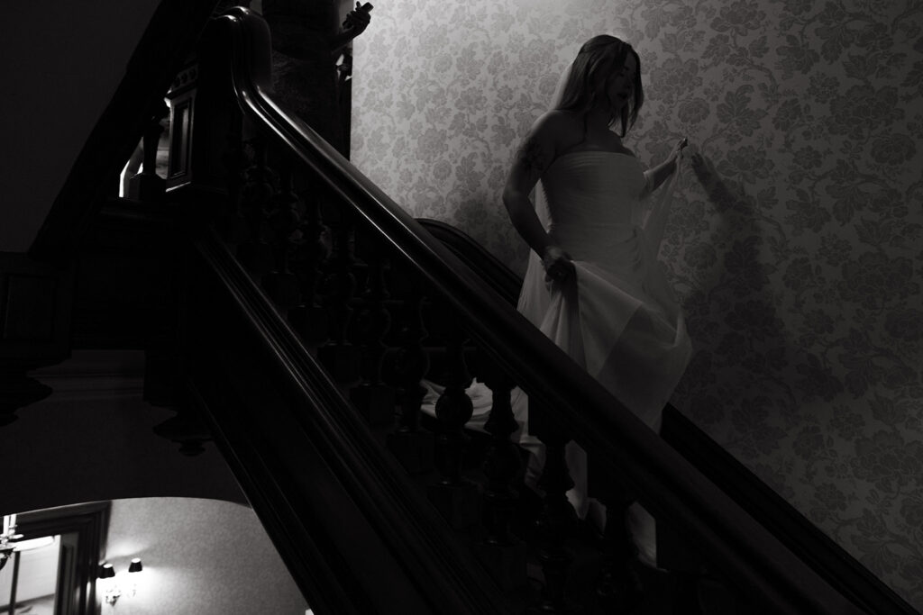 Bride descending the staircase in dramatic black and white, light falling across the bannister.