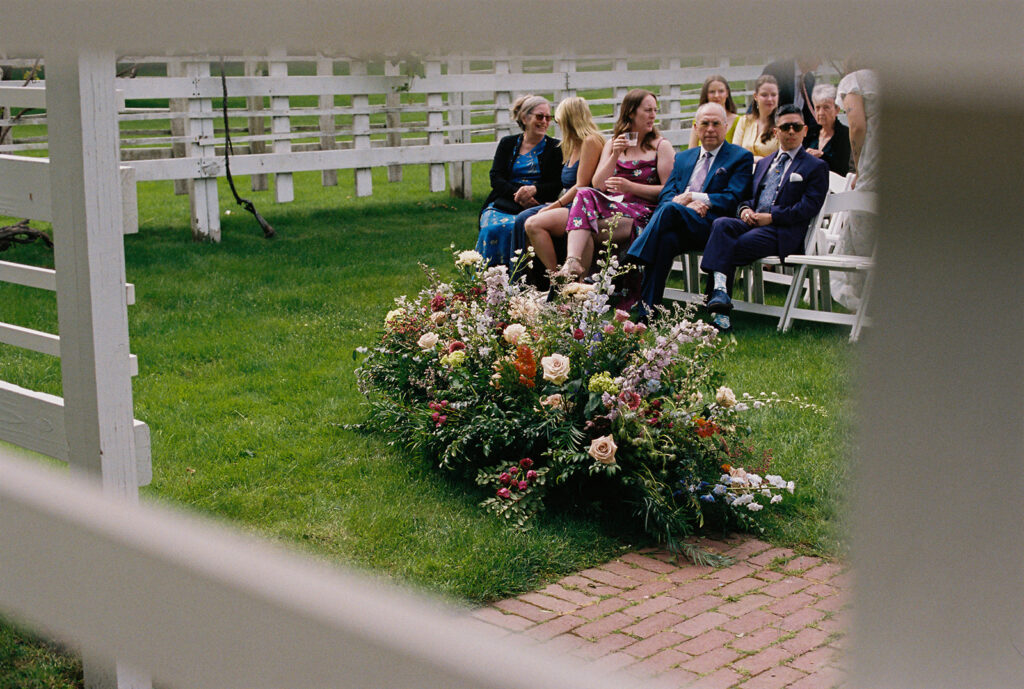 Film wedding photography of ceremony guests seated on white chairs, floral aisle arrangements blooming in the foreground.