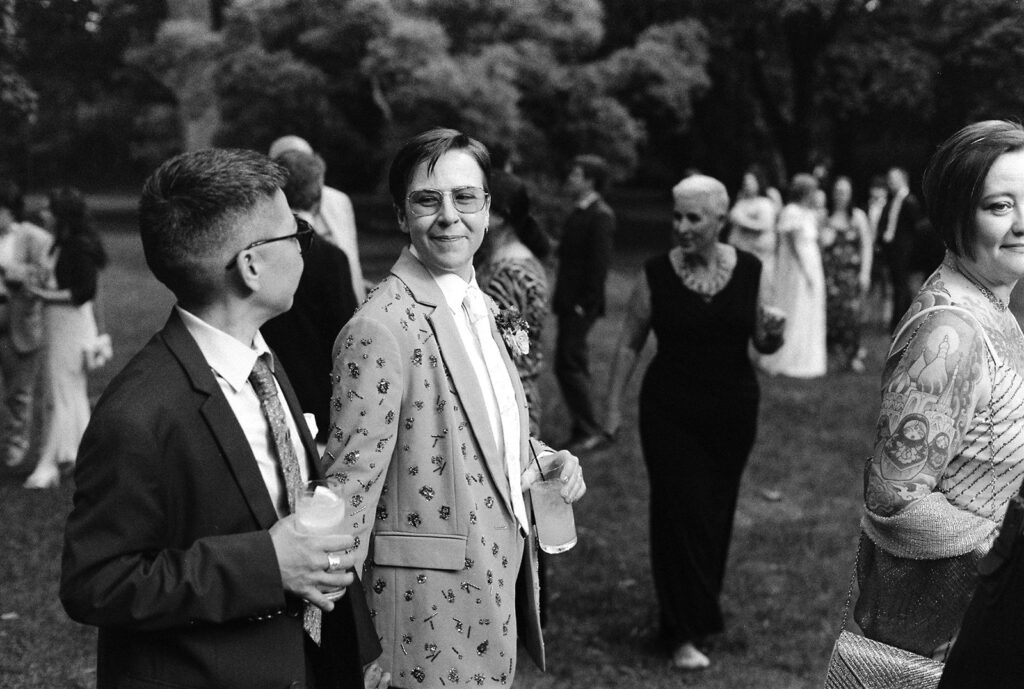 Film wedding photography of guest in patterned suit jacket smiling during cocktail hour, group of guests mingling behind.