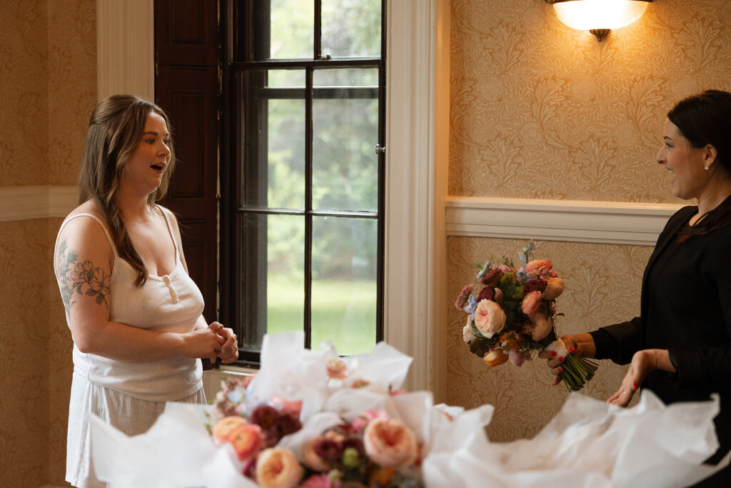 Bride laughing with bridesmaids in golden window light while bouquets are arranged.