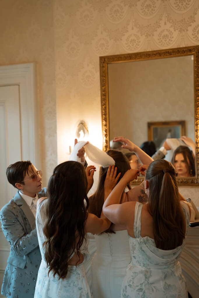 Bridesmaids surrounding bride to adjust her veil, hands working together in soft natural light.