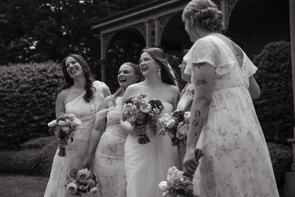 Bridesmaids laughing and holding bouquets outdoors, captured in joyful black and white.