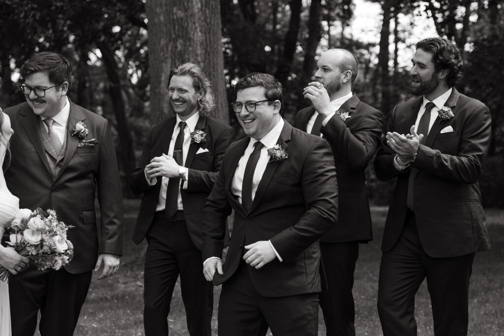 Groom and groomsmen laughing together outdoors, celebratory candid group portrait.