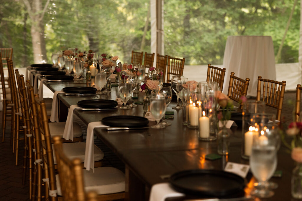 Reception tables under a tent with gold chairs, candles, and floral centerpieces glowing at twilight.