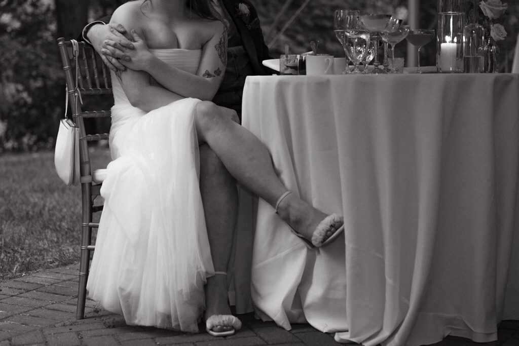 Couple seated at sweetheart table under the reception tent, bride leaning back into groom’s arms.