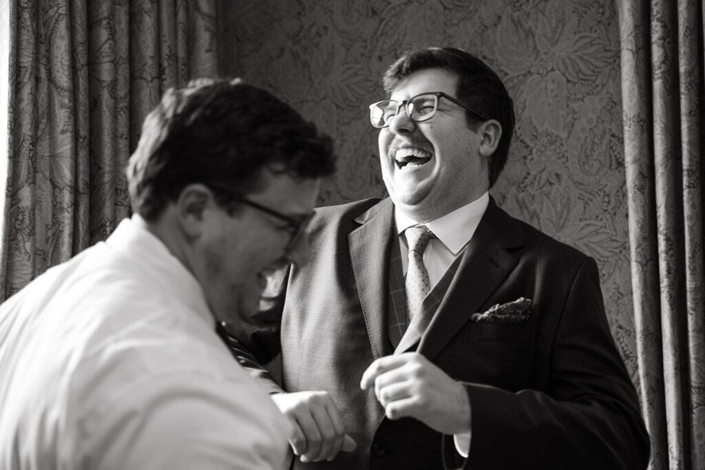 Two groomsmen laughing together while getting dressed, candid black and white film wedding photography moment.