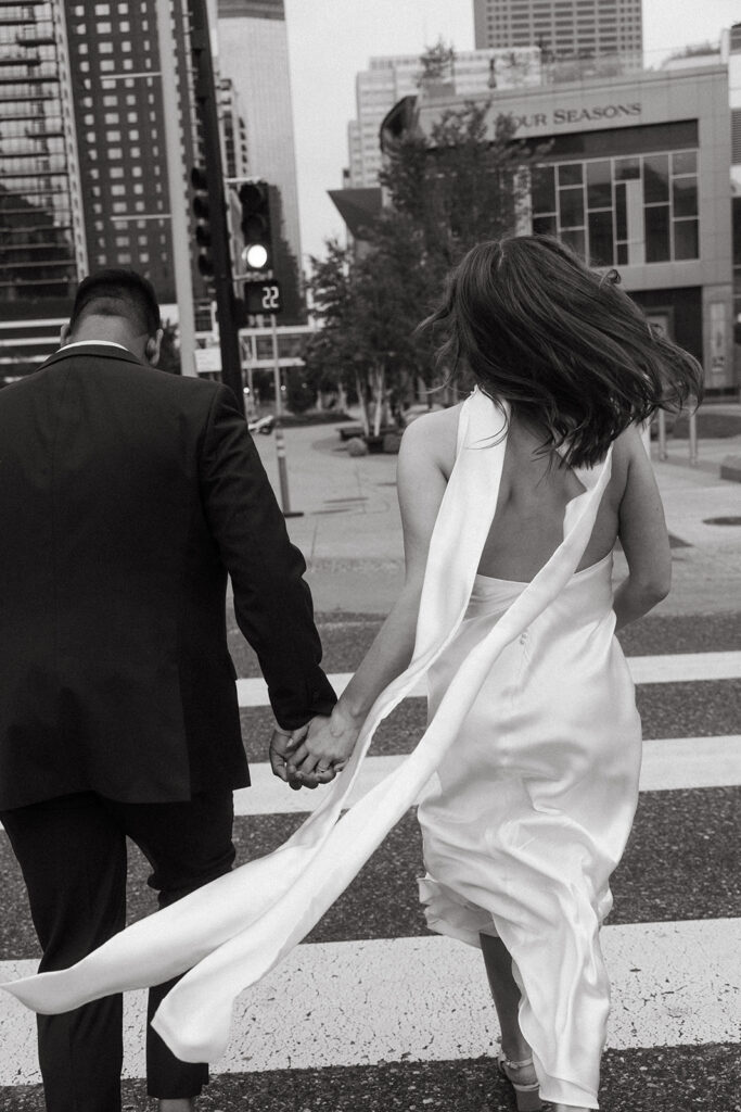 Bride crossing the street in a satin gown with groom beside her, downtown skyline in background.