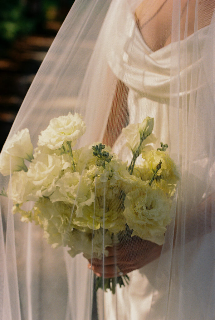 Emma holds her bouquet under her veil as sunlight filters through the fabric for posed wedding photography.