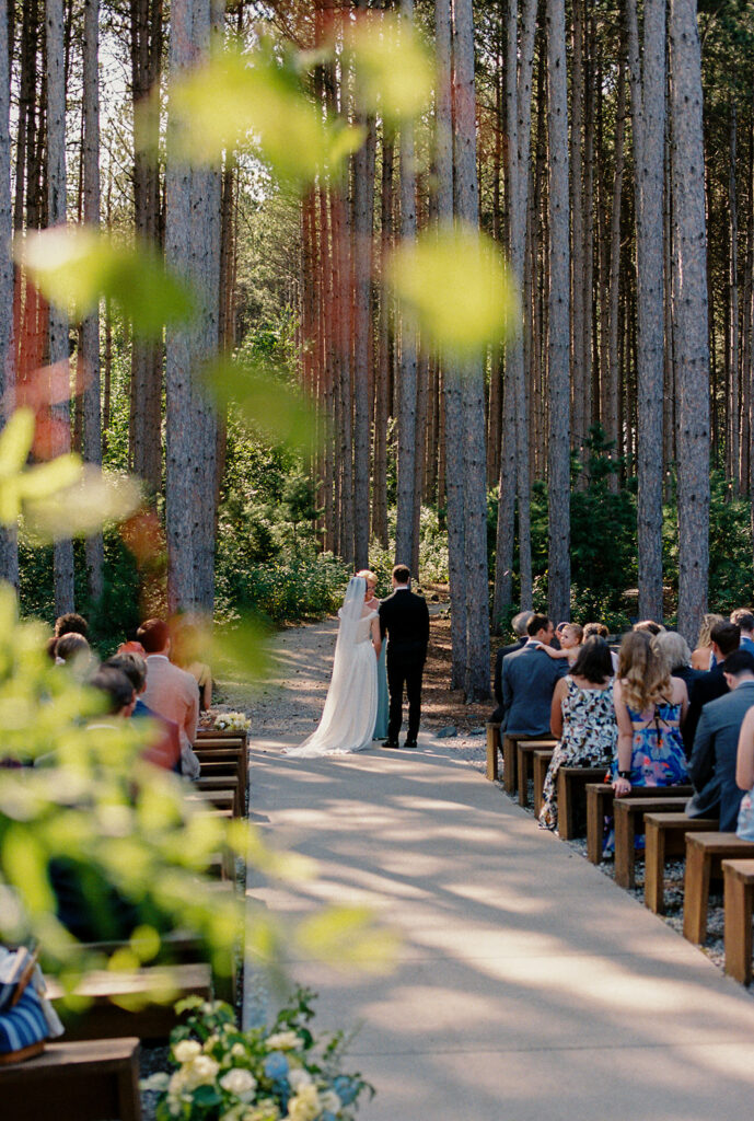 The ceremony unfolds between the pines, every guest stilled by the quiet beauty around them.