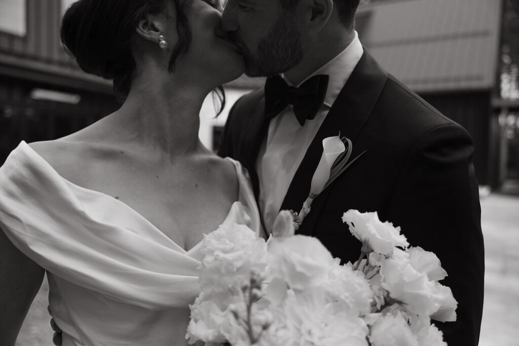 Emma and Isaac kiss outside the venue, her bouquet caught between them, the final frame of a very good story of posed wedding photography.