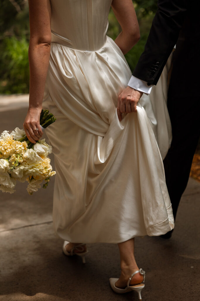 Groom gathering bride's dress while walking they walk down path together in posed wedding photography that feels effortless.