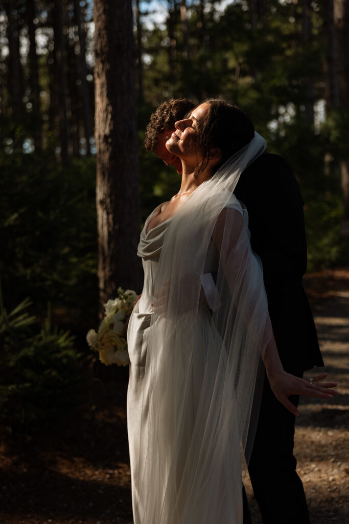 Bride and groom laughing mid-kiss beneath the forest canopy.