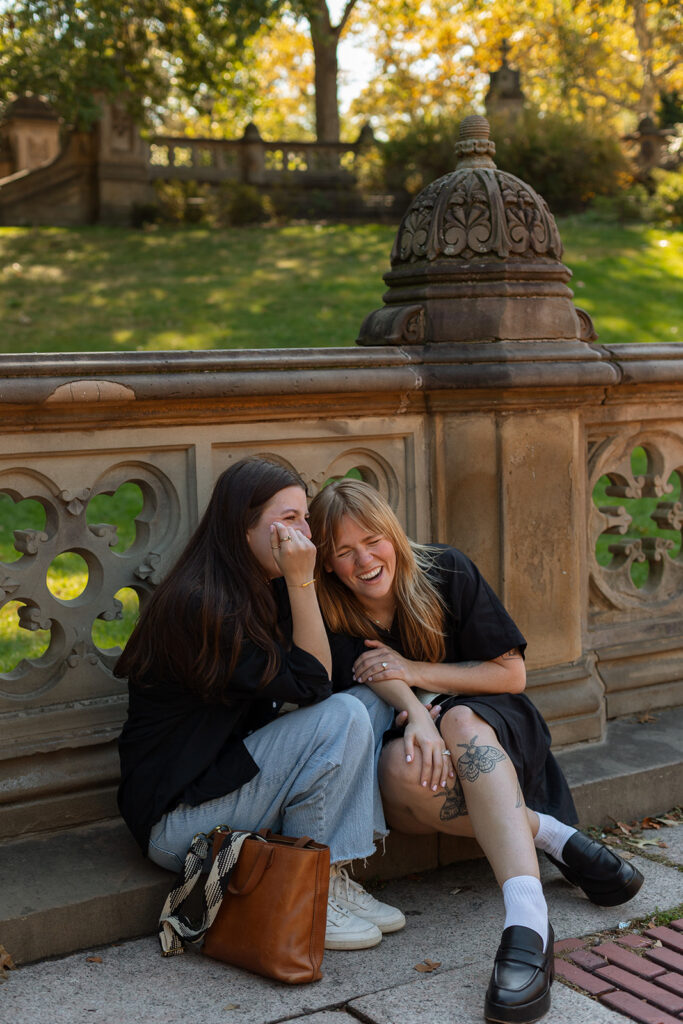A couple laughs with their heads leaned together on the stone terrace in Central Park, surrounded by golden autumn light during their engagement session.