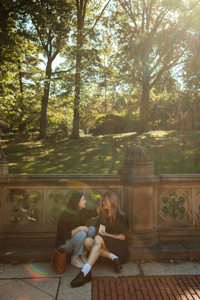 A couple sits against the carved stone railing at Bethesda Terrace, laughing together as sunlight filters through the trees during their New York City wedding proposal.