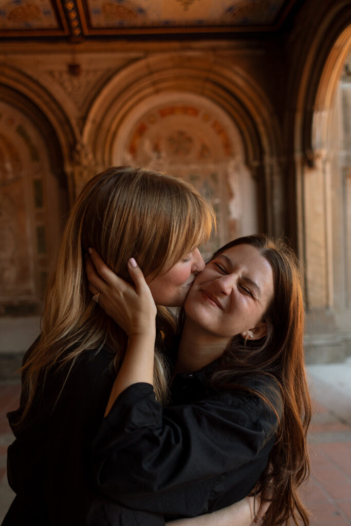 A close-up of one partner kissing the other’s cheek beneath the ornate Bethesda Terrace ceiling during their New York City engagement photos.