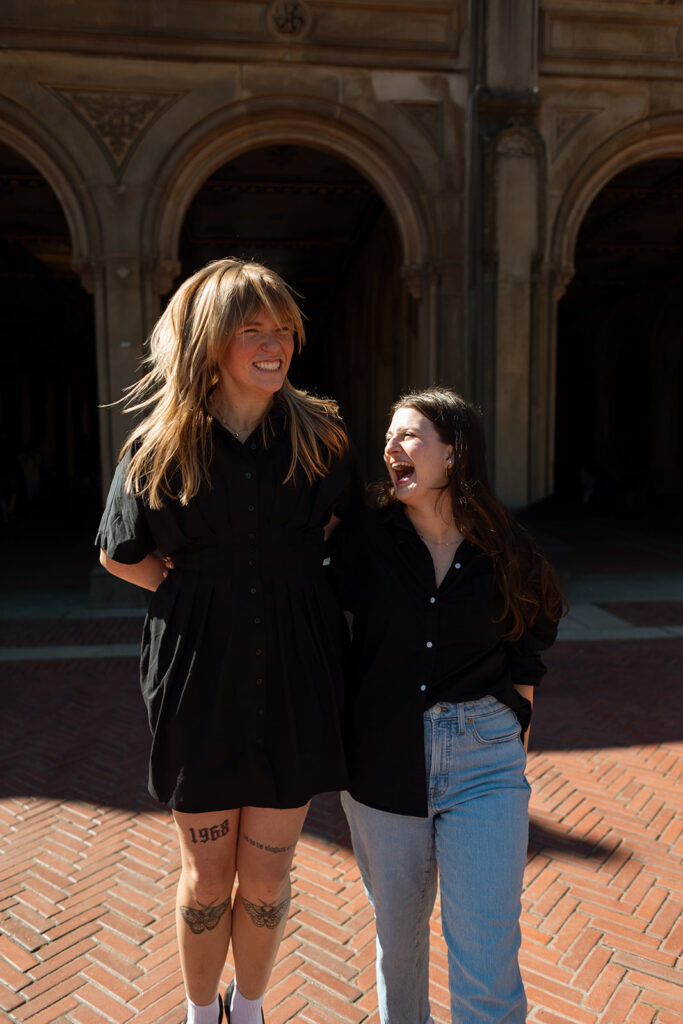 A queer couple walks arm-in-arm under the arches at Bethesda Terrace, sharing a soft smile during their Central Park proposal session.