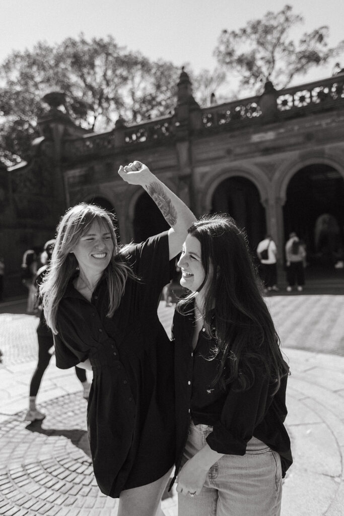 A joyful couple spins each other in the sun outside Bethesda Terrace, laughing during their New York City engagement photoshoot.