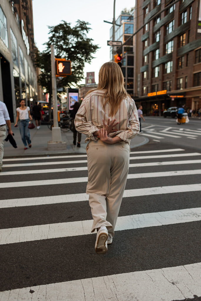 A woman crosses a Manhattan street at sunset, hands clasped behind her back during their New York City engagement session.