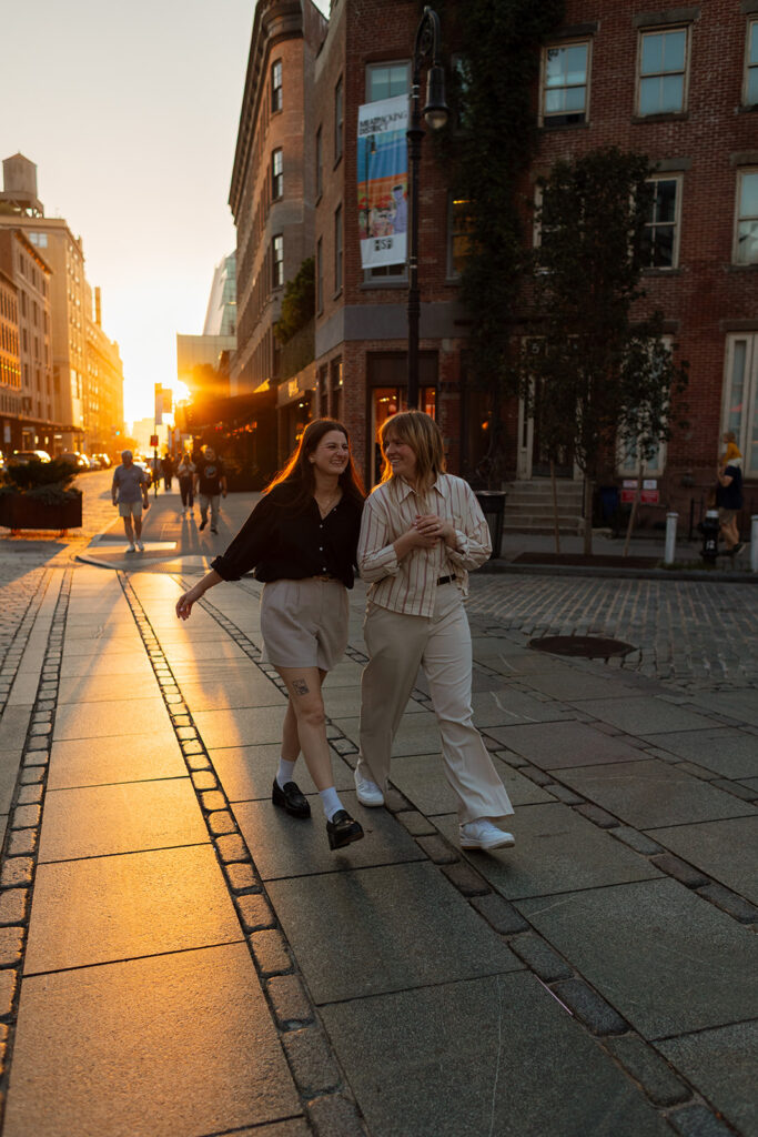 A couple walks arm-in-arm down a cobblestone street at golden hour, laughing as the city glows behind them during their NYC engagement photos.