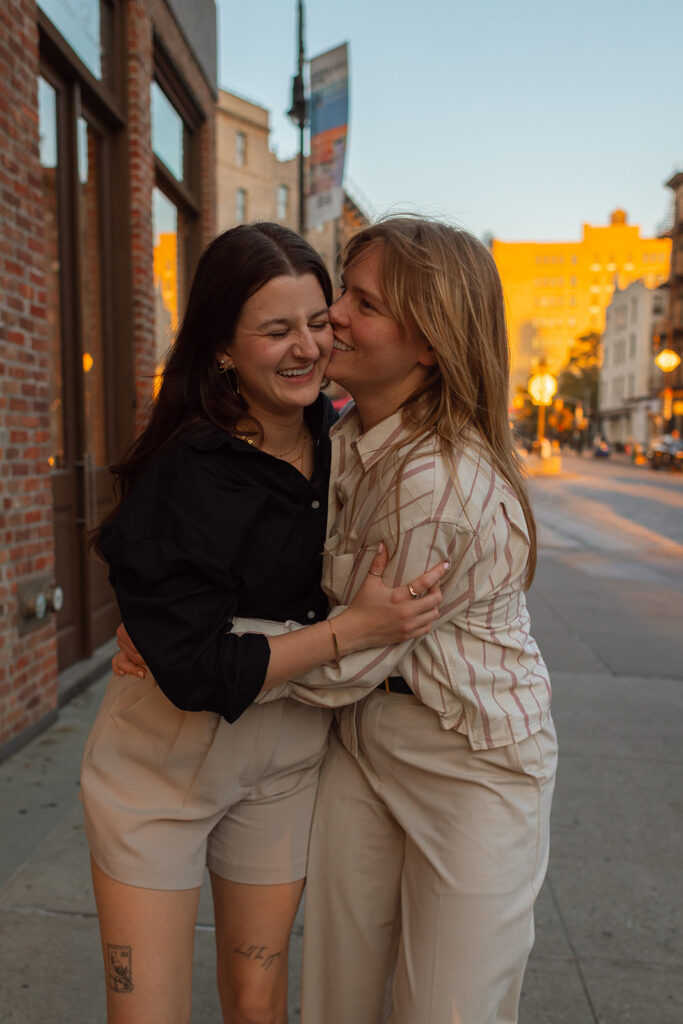 A couple laughs and embraces on a New York sidewalk at sunset, holding each other during their intimate LGBTQ+ engagement session.