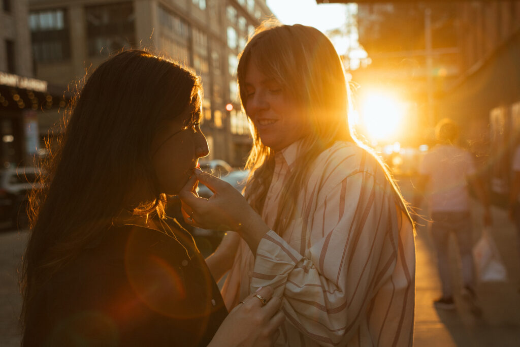 A queer couple stands close in a New York City street at golden hour, sharing a joint as warm sunlight and lens flare wrap around them during their documentary-style engagement photos.
