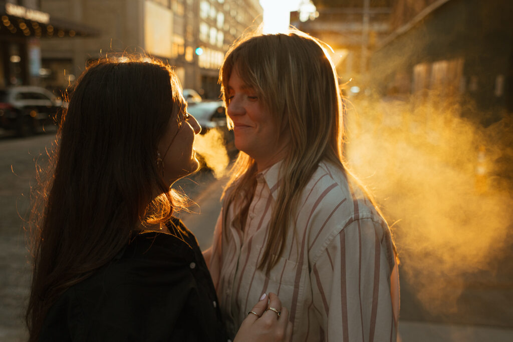 A woman exhales smoke into the golden-hour glow while her partner smiles beside her, the haze catching the evening light during their New York city wedding proposal engagement photos.