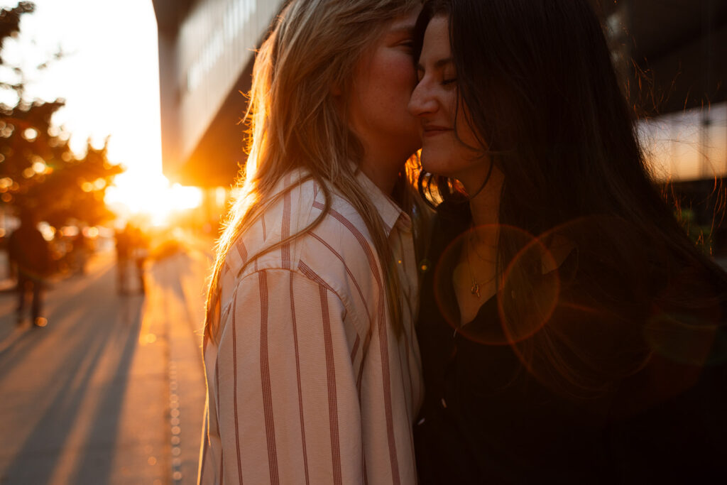 A queer couple kisses at golden hour in Manhattan, warm sunlight and lens flare wrapping around them during documentary-style proposal photos.