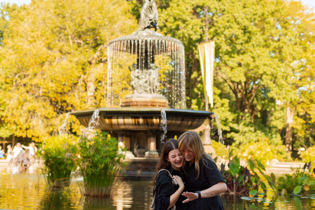 A newly engaged queer couple hugs in front of the Angel of the Waters fountain, golden afternoon light pouring over them during their New York City wedding proposal photos in Central Park.