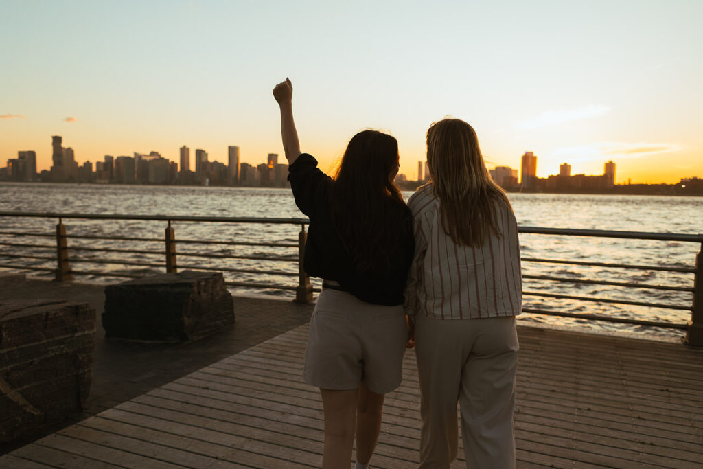 A couple raises an arm toward the New York skyline at dusk, celebrating their engagement along the Hudson River during their documentary-style photoshoot.