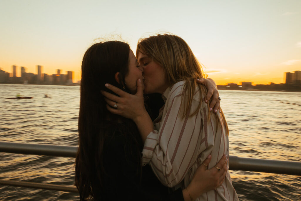 A queer couple kisses at the Hudson River waterfront at sunset, holding each other close during their NYC engagement photoshoot.