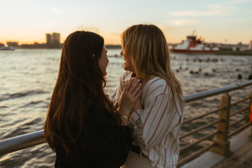 A couple stands at the Hudson River waterfront at sunset, leaning in close and sharing a quiet moment during their New York City engagement photos.