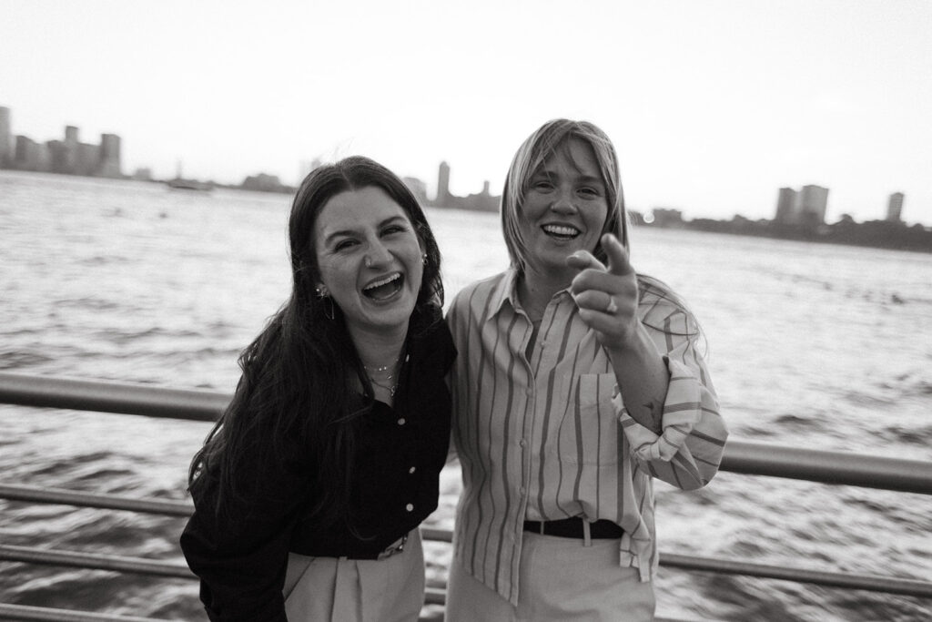 A black-and-white photo of a queer couple laughing together along the Hudson River, the skyline behind them during their NYC proposal session.