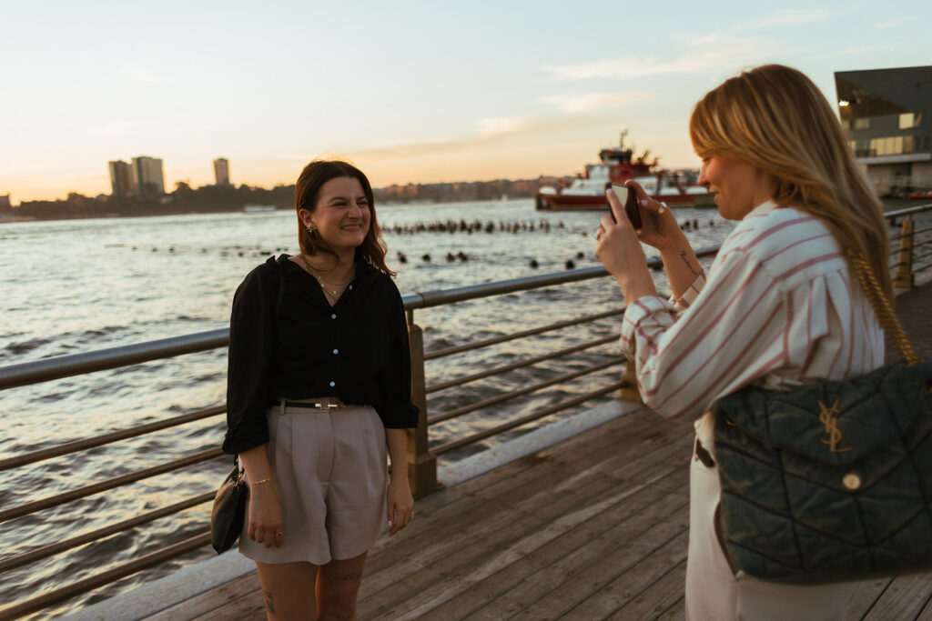 One partner photographs the other along the Hudson River boardwalk as the sun sets behind the city during their engagement session.