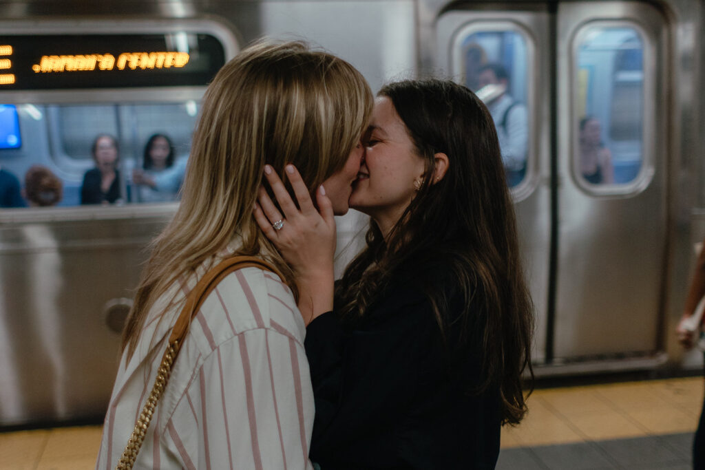 A queer couple kisses on the subway platform as a train idles behind them, surrounded by commuters during their candid NYC engagement photos.