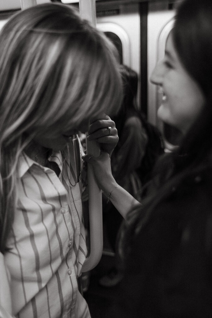 A woman gently holds her partner’s hand while gripping the subway pole, sharing a soft smile on a packed New York City train.