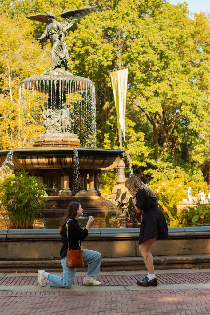 A woman kneels to propose to her partner in front of the Angel of the Waters fountain at Bethesda Terrace in Central Park during a heartfelt LGBTQ+ New York City proposal.