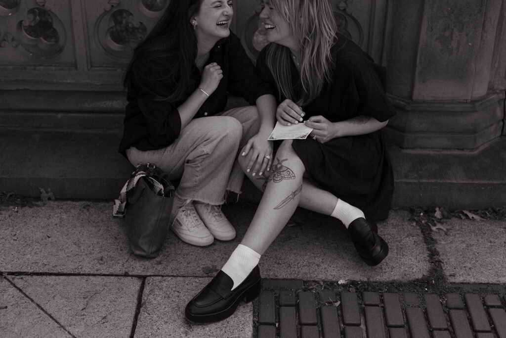 Two women sit on the ground in Central Park, laughing together with their knees touching, holding handwritten proposal letters during an NYC LGBTQ+ engagement session.