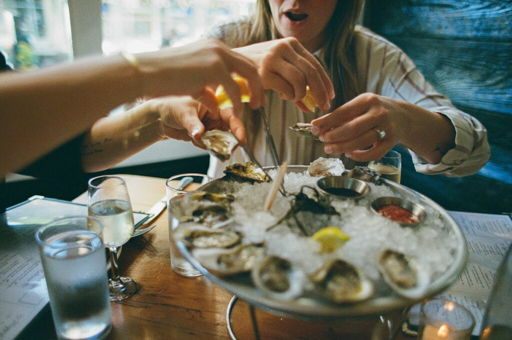A couple reaches for oysters on ice at a New York City restaurant, celebrating their engagement with a joyful, candid moment.