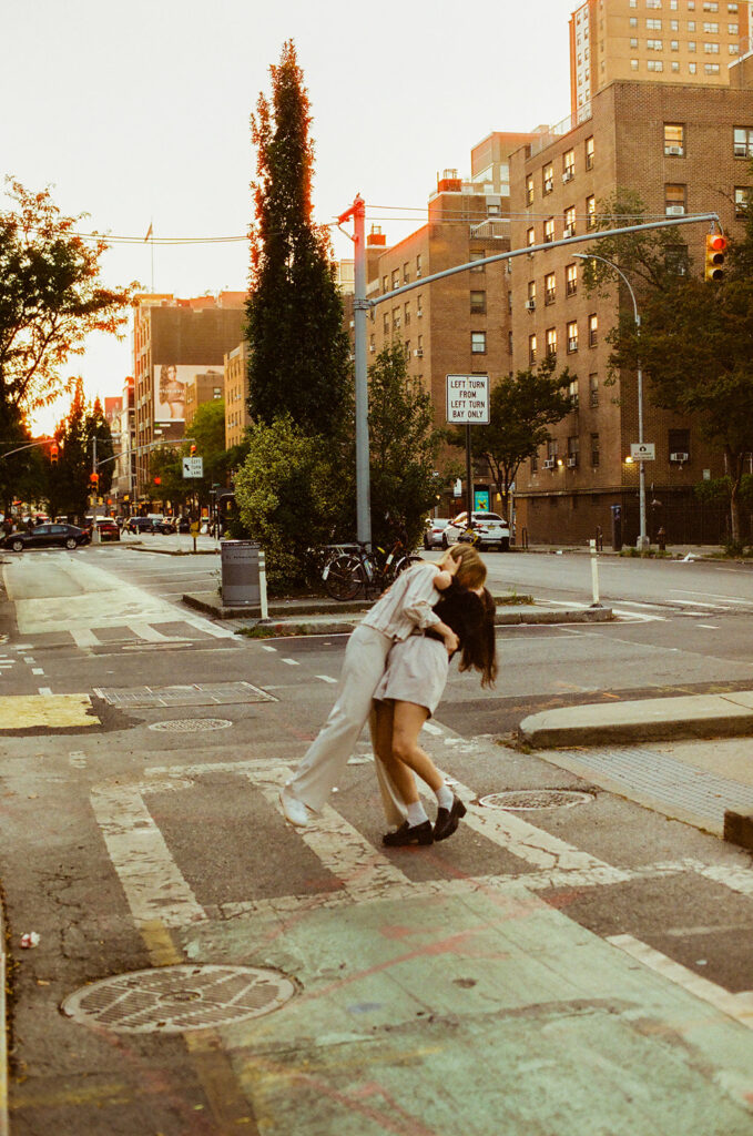 A couple shares a dramatic dip and kiss in a Manhattan crosswalk at sunset, the city glowing behind them during their New York proposal session.