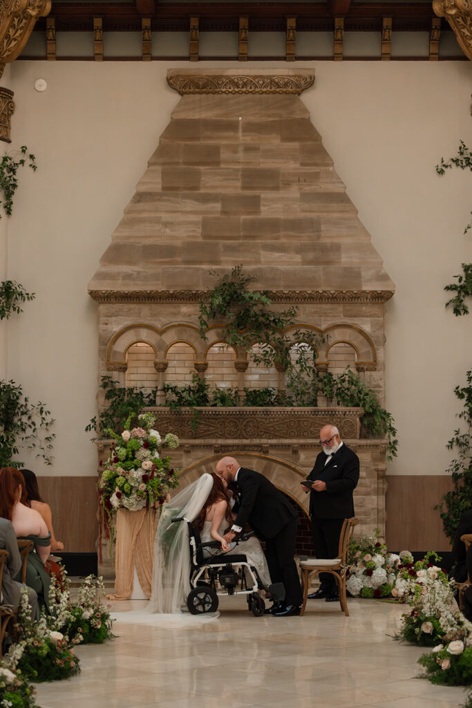 Groom bends down to kiss his bride in her wheelchair during their low-light wedding at Union Station in Nashville.
