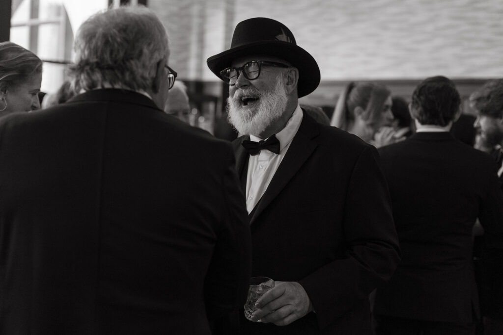 Father of the bride wearing top hat and glasses laughs with sheer abandon at something a wedding guest said during cocktail hour