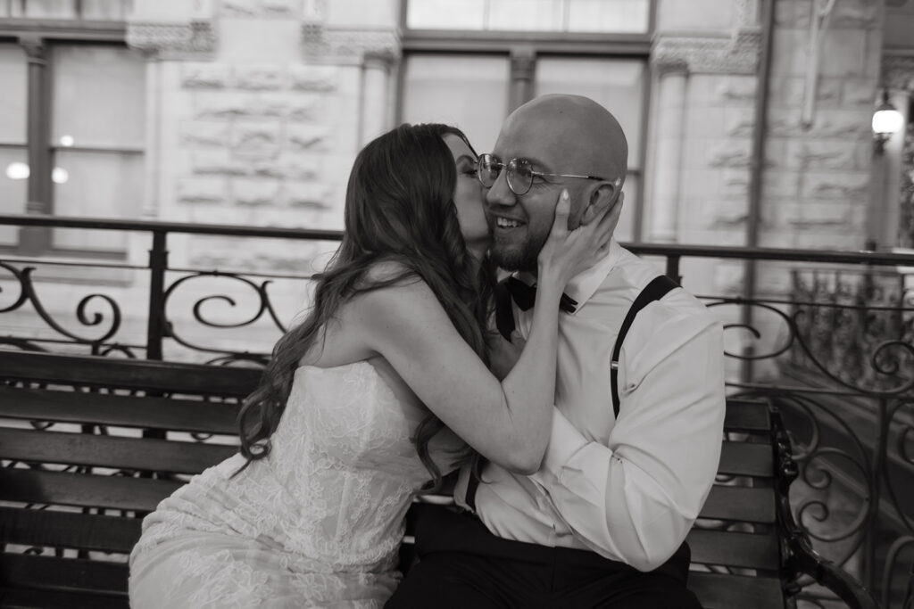 Bride grabs her husband's face and kisses his cheek as they sit on park bench outside Union Station in Nashville