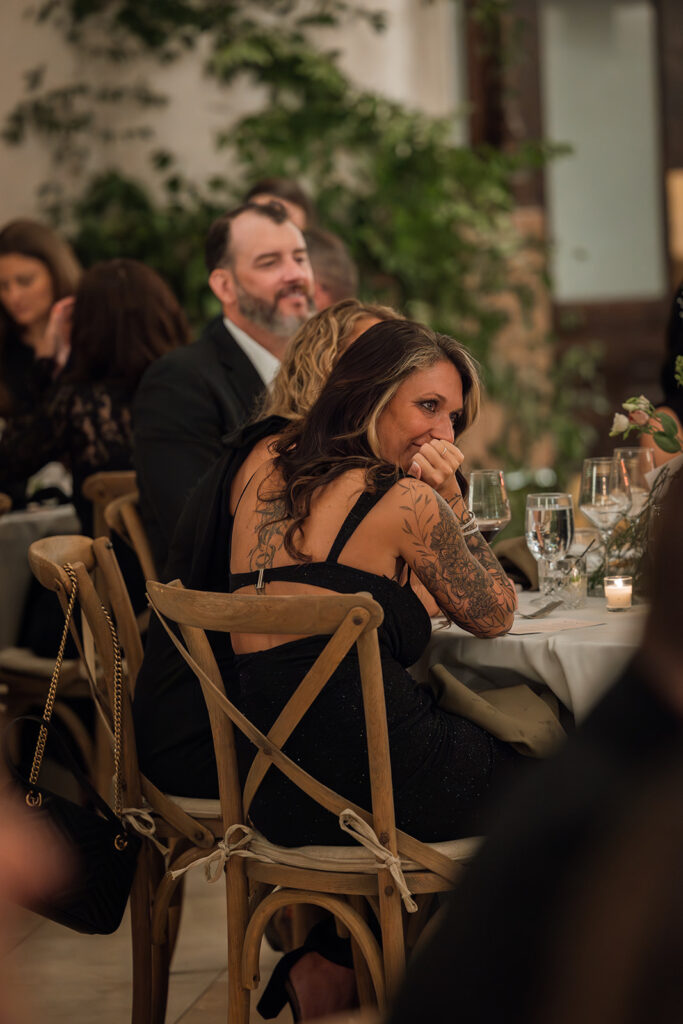 A wedding guest with sleeve tattoos rests her head in her hands as she listens to emotional speech