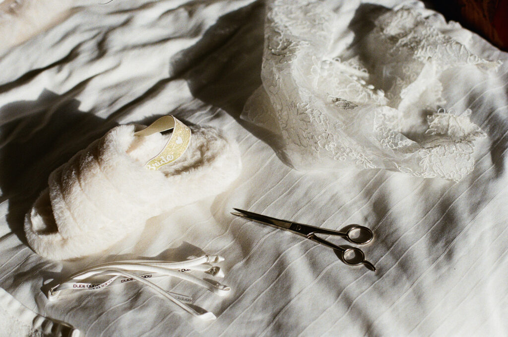 Film still of bride's dress, slippers, and a pair of sewing scissors resting on white linen sheets.