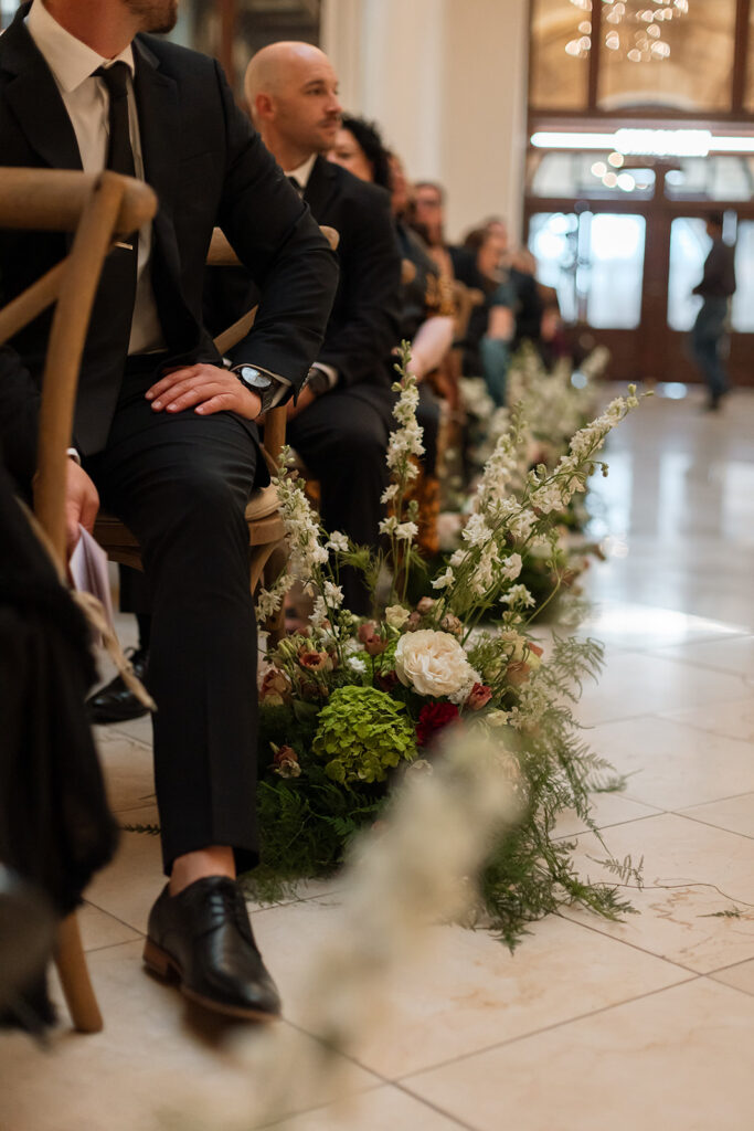 Guests' hand rests on his leg as he turns to see bride make her way down the aisle.