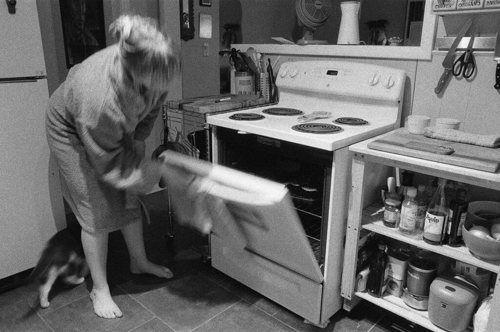 Black-and-white documentary photograph of Nashville photographer Ali Miller opening the oven while baking with her cat nearby.
