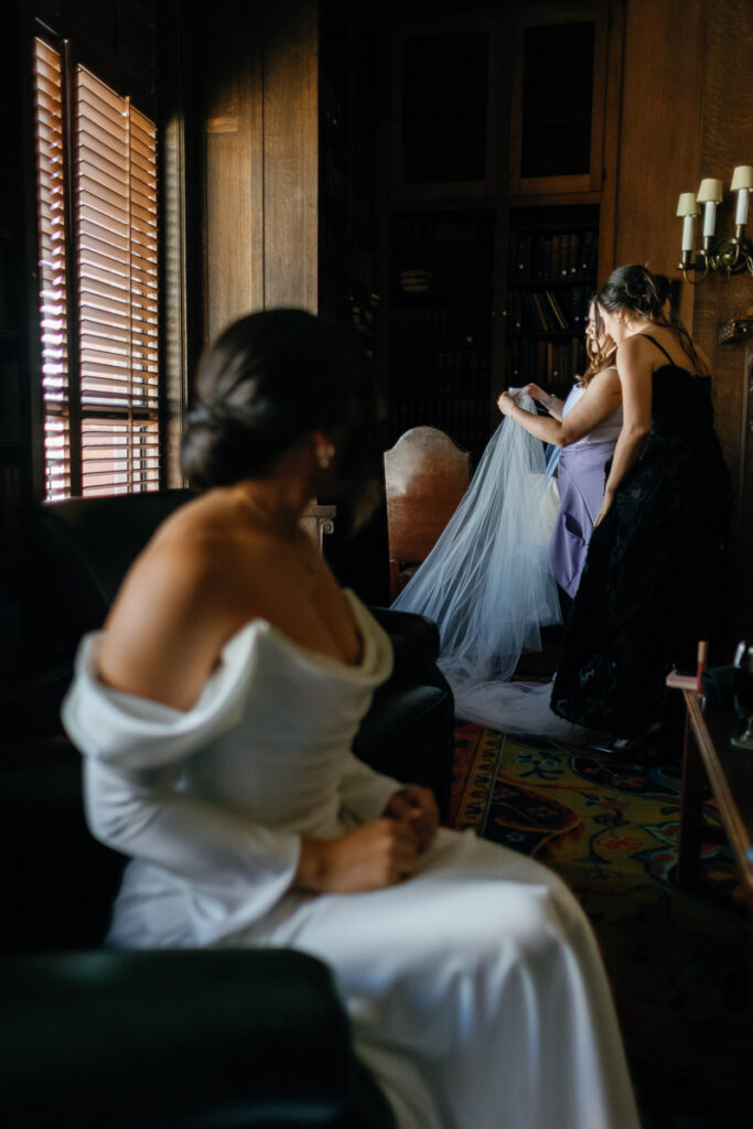 Bride looks on as other bride gets her wedding veil placed in candid wedding photo