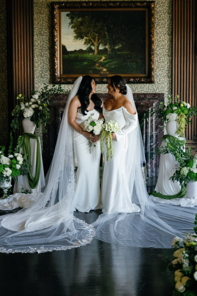 Lesbian brides pose for portrait in front of stunning wood fireplace with beautiful painting and flowers made by a friend surrounding them
