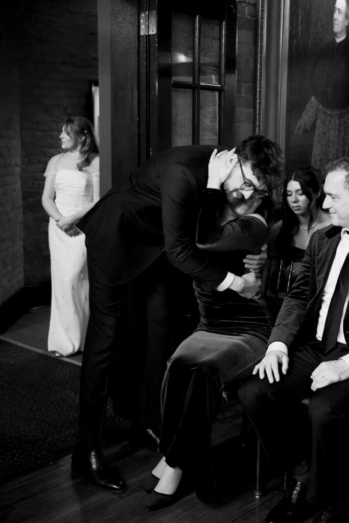 Groom leans over to hug his mom as bride stands in the hallway in the background, proof that meaningful wedding ideas start with being fully present